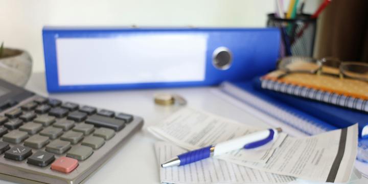 A blue lever-arch file, spiral notebooks, a desk calculator and a pen lying on printed slips on a white desk.