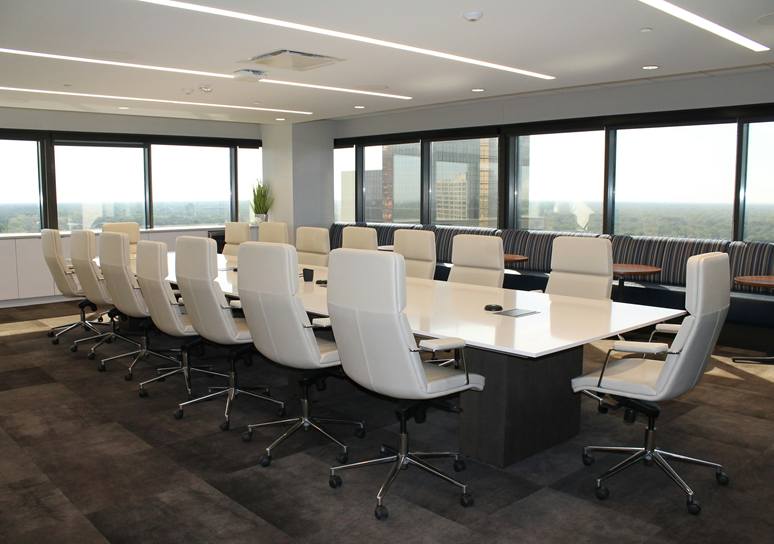 An empty boardroom with a long white table, white high-back chairs and windows on two sides overlooking office towers and treetops.