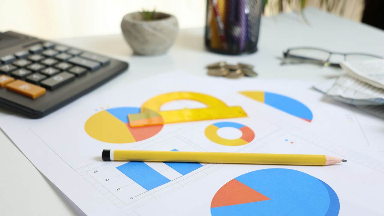 A calculator, a pencil, a yellow protractor and printed pie and bar charts lie on a white desk, with coins, glasses, a pen holder and a small plant pot out of focus behind.
