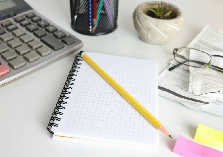 A desk calculator, a squared spiral notebook with a yellow pencil, a pen holder and reading glasses resting on printed receipts on a white desk.