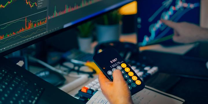 At a dimly lit desk with monitors showing candlestick charts, one hand holds a phone calculator above a notebook while another hand points at a screen.
