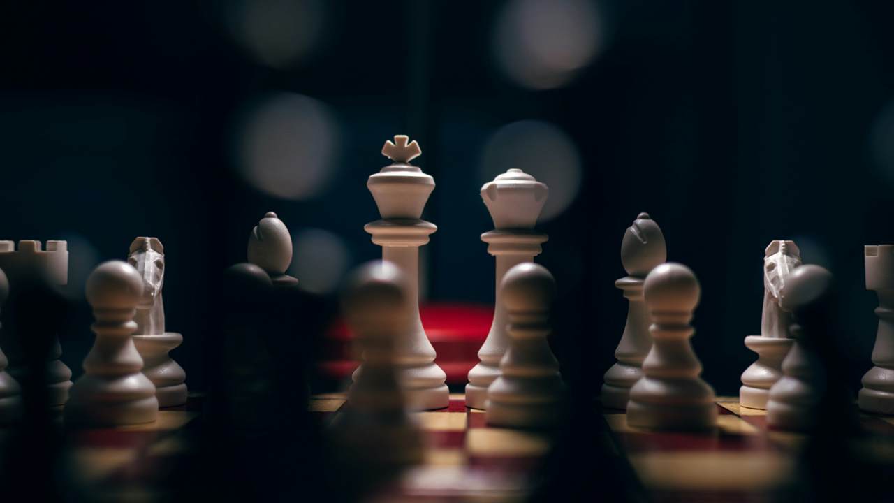 A white chess king and queen stand in sharp focus behind a row of blurred pawns, against a dark blue background.