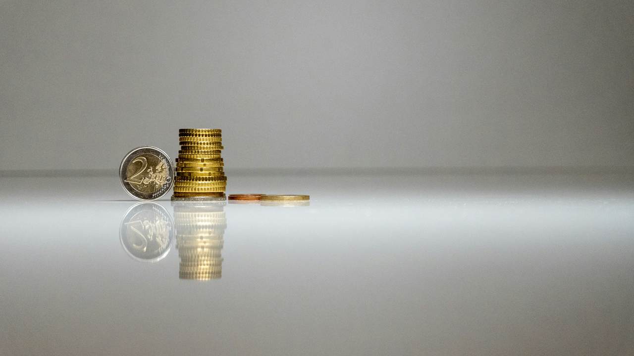 A stack of gold coloured coins with a larger coin leaning against it and two coins lying flat beside it, reflected in a glossy grey surface.
