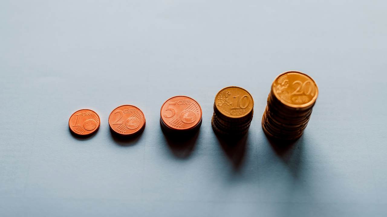 Copper and gold coloured coins arranged in a row on a pale blue-grey surface, rising from a single coin on the left to a tall stack on the right.