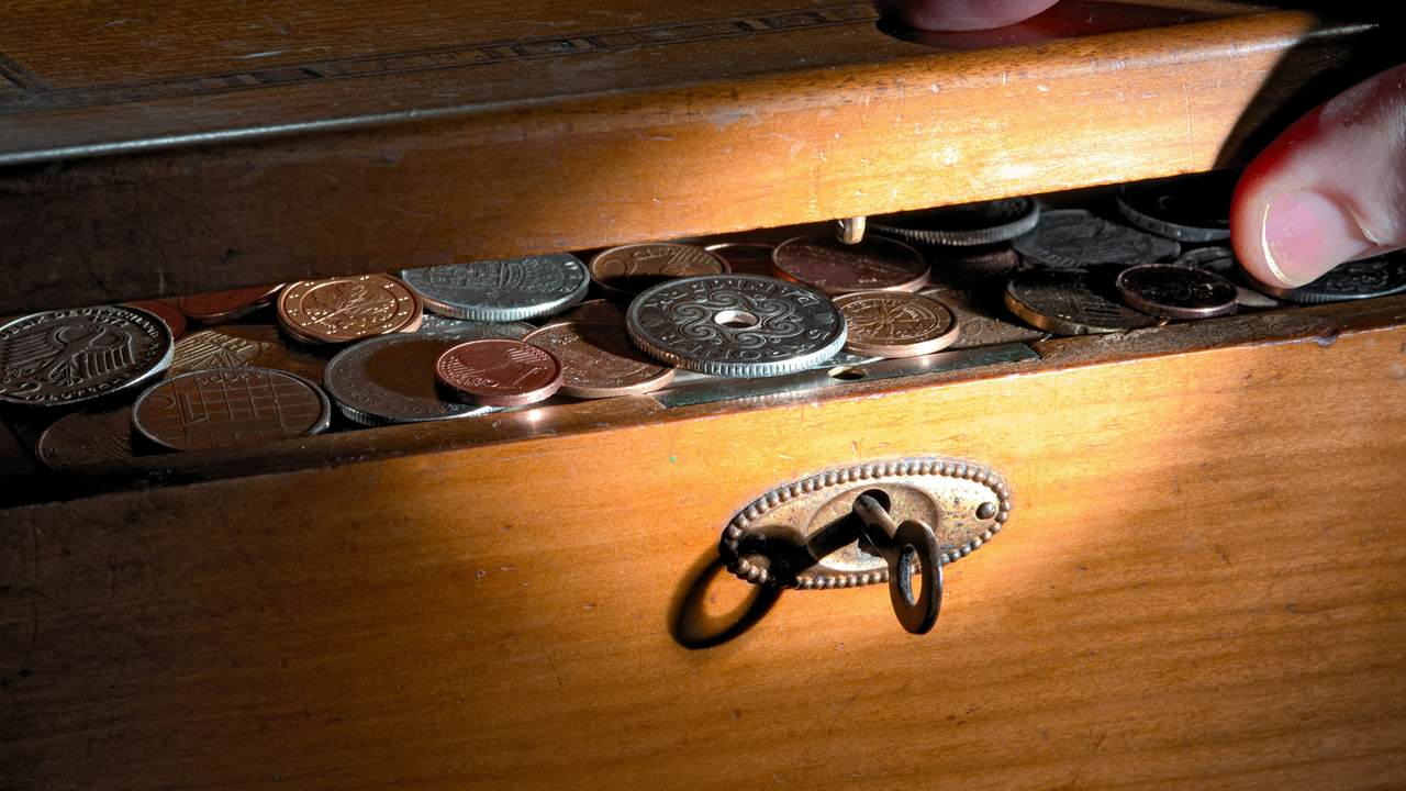 A hand pulls open a wooden drawer filled with mixed coins, with a small key in the lock on the drawer front.
