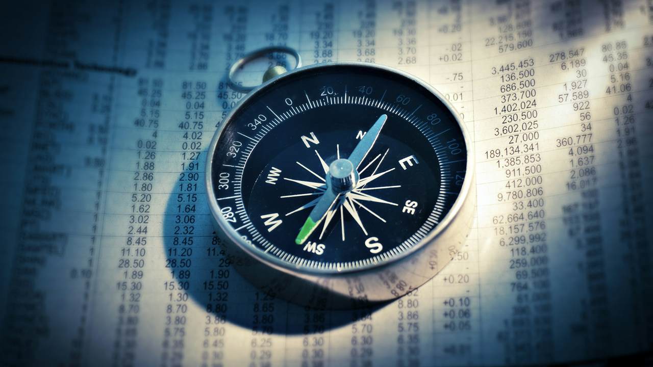 A compass lies on a printed table of share prices, in blue-tinted light.