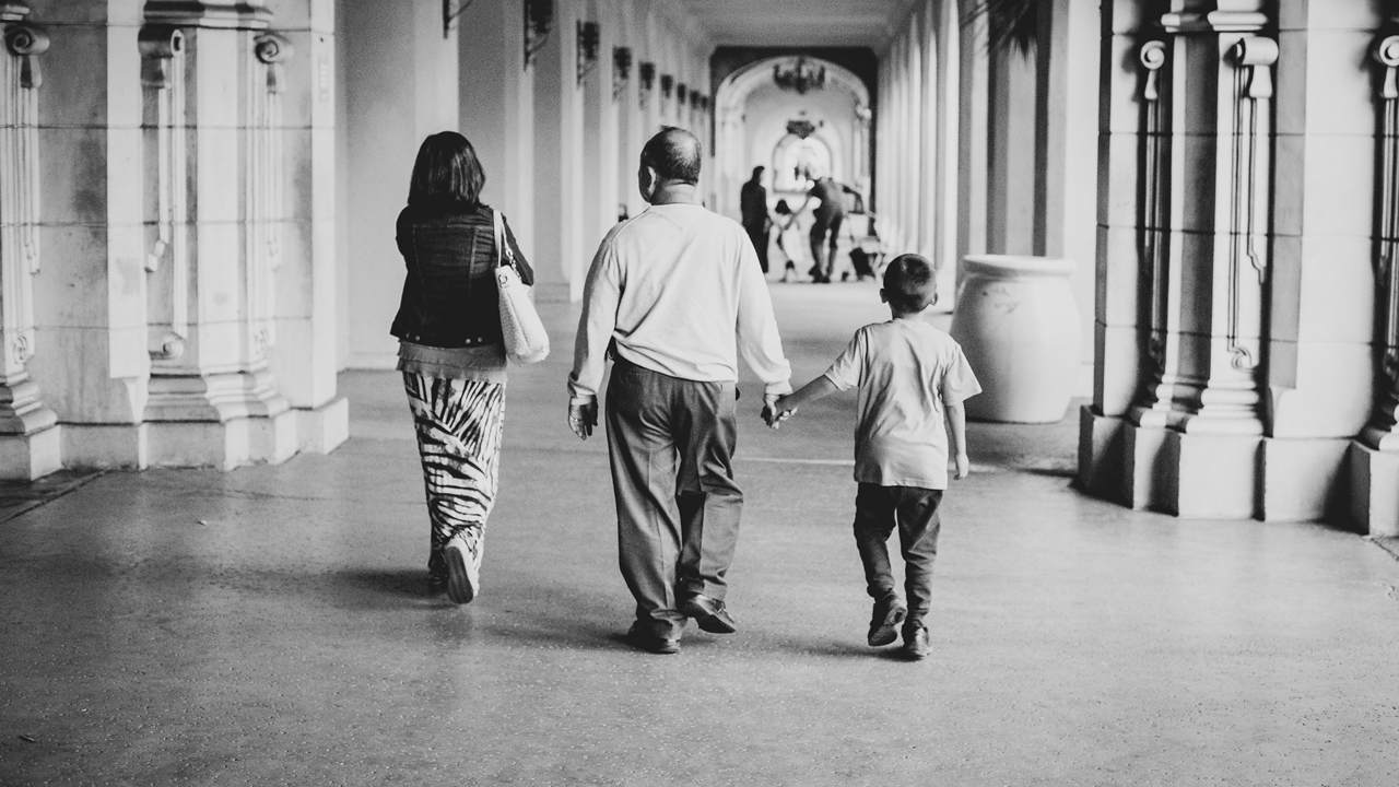 Black and white photo, seen from behind, of a woman, an older man and a boy walking along a covered arcade; the boy holds the man's hand.