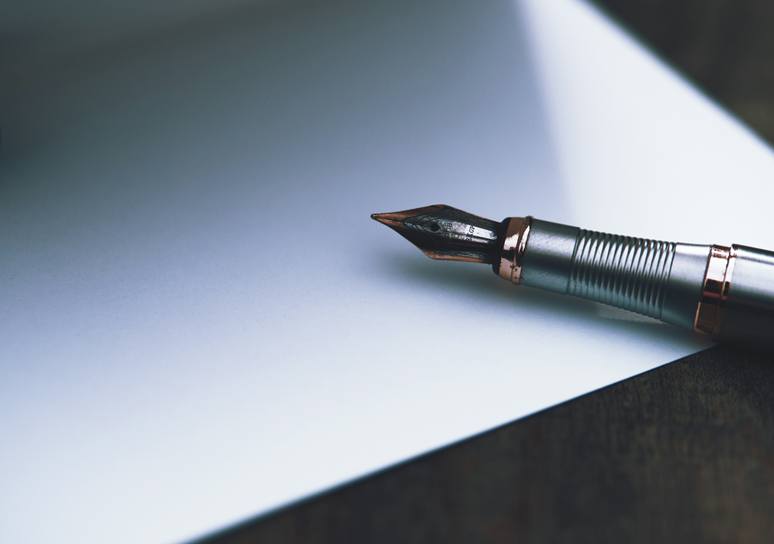 A silver fountain pen with copper-coloured trim rests on a blank sheet of paper on a dark wooden desk.