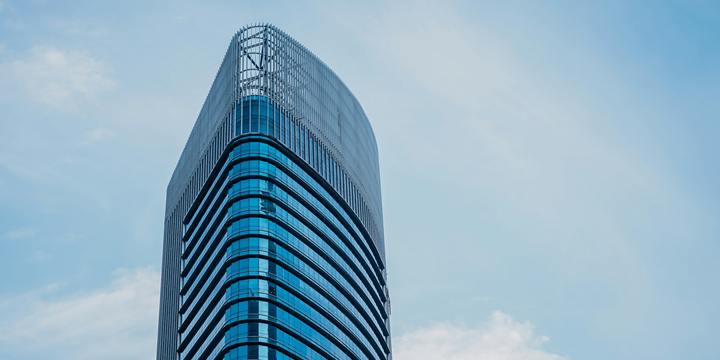 A tall glass office tower with a rounded crown against a soft blue sky.