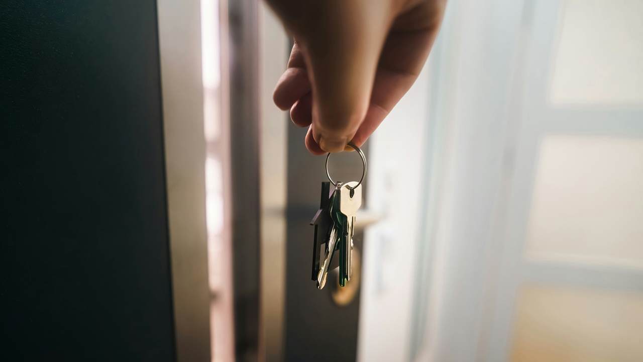 A hand holds a ring of keys with a small house-shaped fob in front of a dark door that stands slightly open.