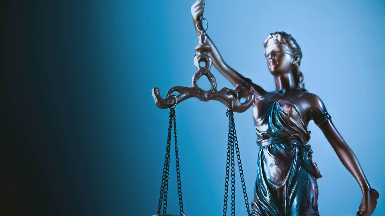 A bronze coloured statuette of blindfolded Lady Justice holds up a pair of scales against a blue background.