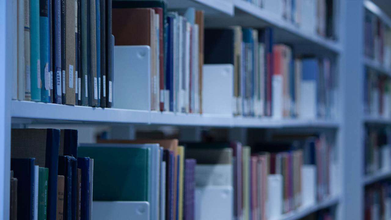 Deretan buku dan kotak arsip putih di rak perpustakaan logam, difoto dalam cahaya kebiruan dengan bagian ujung yang tampak buram.