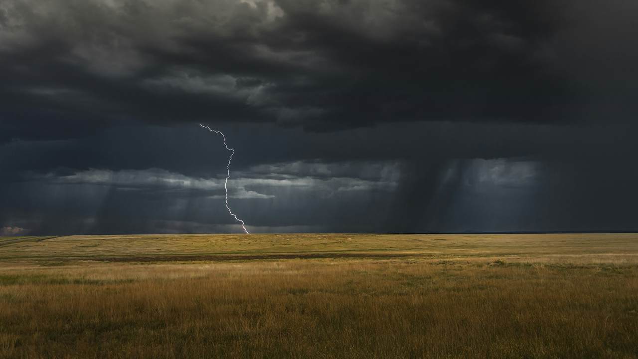 A bolt of lightning strikes a flat grassy plain beneath heavy dark storm clouds and distant curtains of rain.