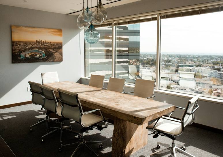 A sunlit meeting room with a solid timber table, white chairs, blown-glass pendant lamps, a framed city photograph and a wide window overlooking the city.