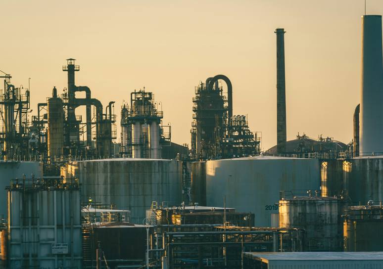 Distillation columns, chimneys and storage tanks of an oil refinery outlined against a warm golden sky.
