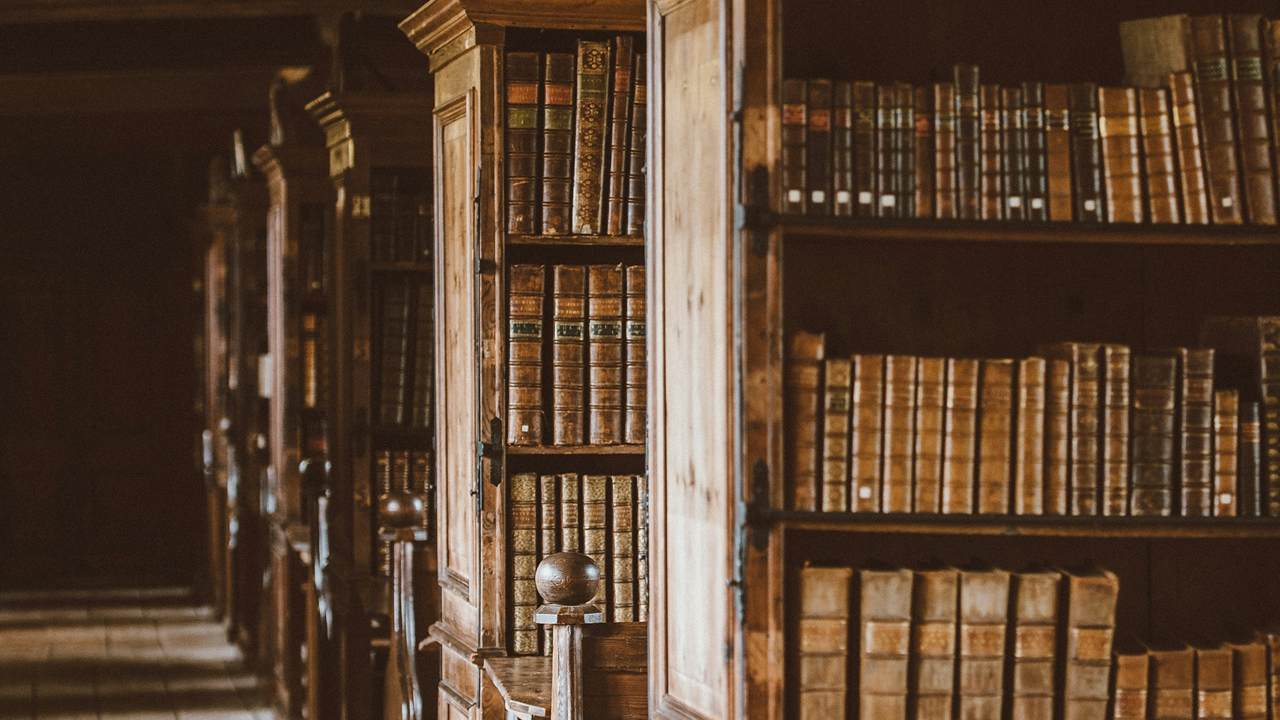 Buku-buku bersampul kulit memenuhi lemari kayu tinggi di sepanjang lorong sebuah perpustakaan tua.