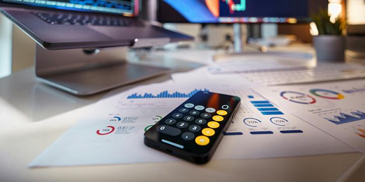 A phone showing a calculator rests on printed charts on a desk, with a laptop and a monitor displaying price charts out of focus behind it.