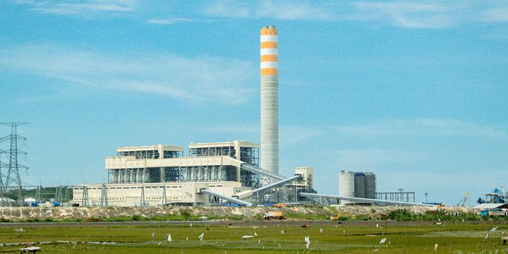 A thermal power station with a tall orange and white striped chimney and conveyor galleries rises behind green rice fields under a blue sky.