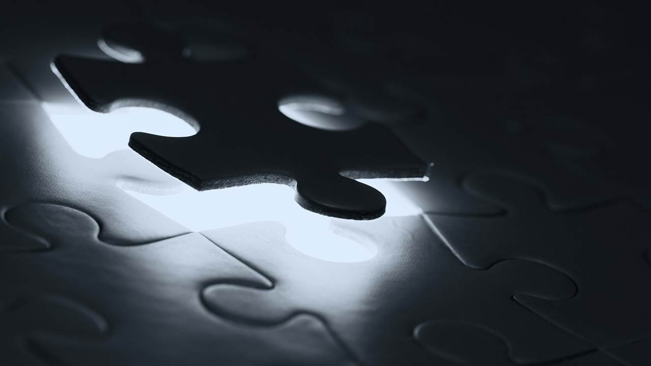A dark jigsaw piece hovers above the matching gap in a puzzle, with light shining up through the opening.