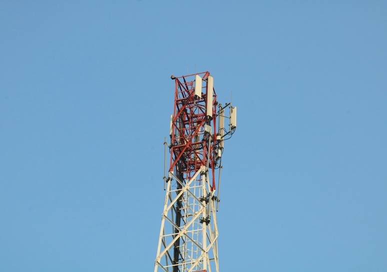 The top of a red and white lattice telecommunications tower carrying mobile network antennas against a clear blue sky.