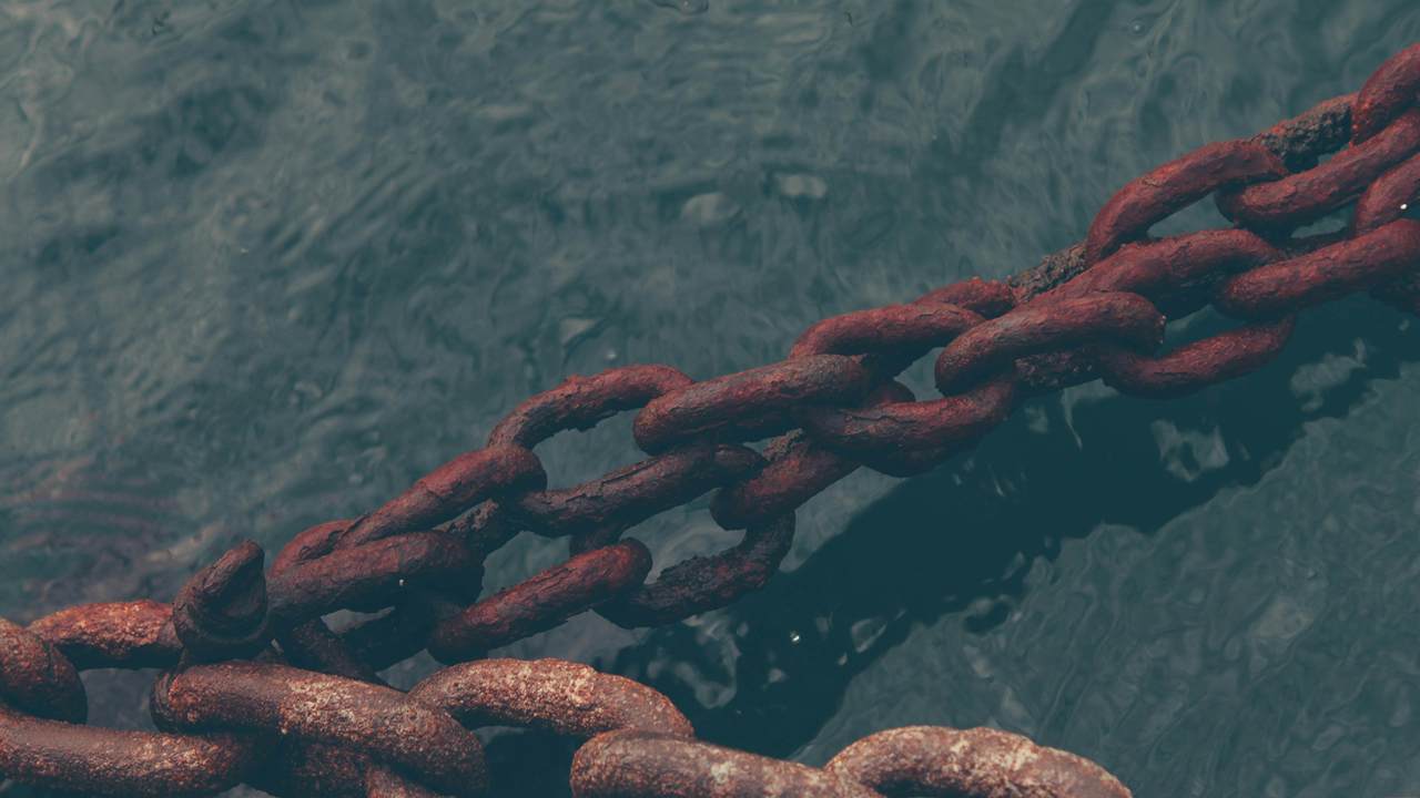 Rantai besar yang berkarat membentang miring di atas air berwarna hijau kebiruan yang gelap.