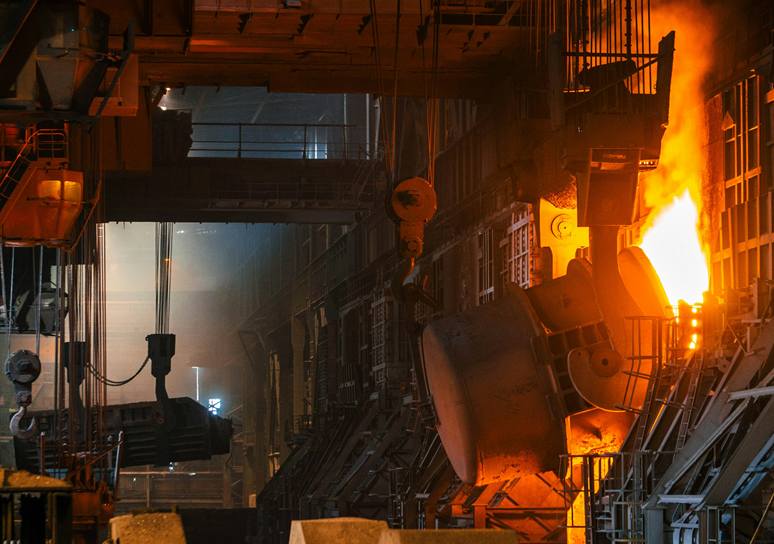 Inside a steel mill, a large ladle tilts beside a blaze of molten metal while crane hooks hang in the smoky hall.