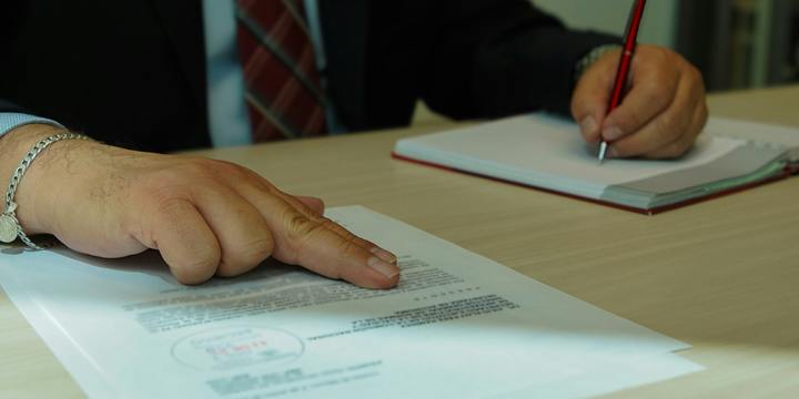 A man in a dark suit and striped tie rests two fingers on a stamped printed document while writing in a notebook with a red pen.