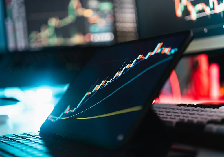 A tablet on a desk shows a rising candlestick chart with trend lines, surrounded by blurred trading monitors and a keyboard.