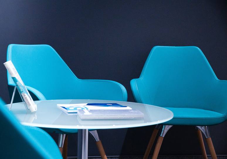 Two turquoise armchairs and a round glass table with a few brochures stand against a dark navy wall.