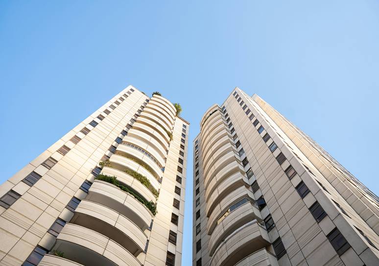 Two beige high-rise apartment towers with curved balconies and roof gardens, seen from below against a blue sky.