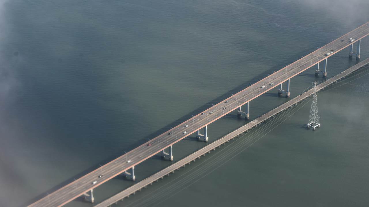 Pemandangan dari udara sebuah jembatan jalan raya panjang dan jembatan sejajar yang lebih sempit melintasi perairan tenang berwarna hijau keabu-abuan, dengan kabut tipis di tepinya.