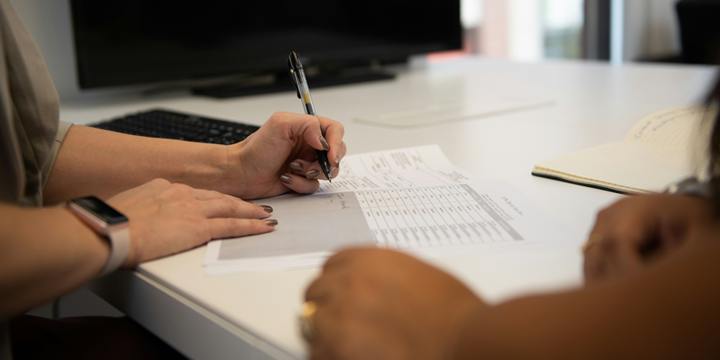 Two people sit at a white desk as one writes on a printed worksheet with a pen, with an open notebook and a monitor nearby.