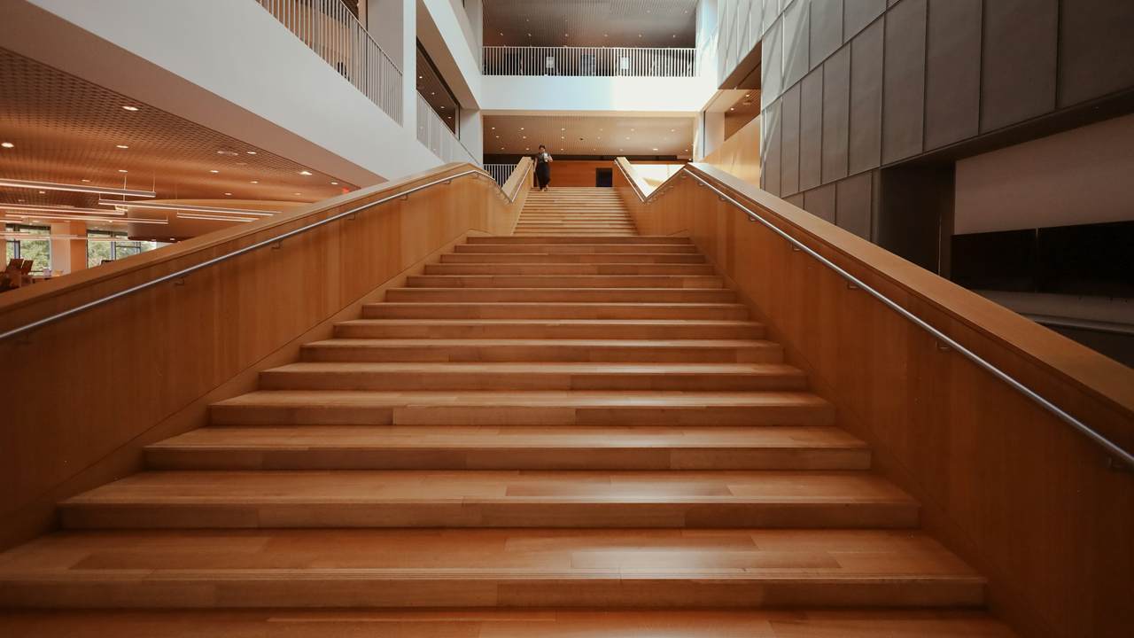 Tangga kayu lebar dengan pegangan baja menanjak di tengah atrium modern yang terang.
